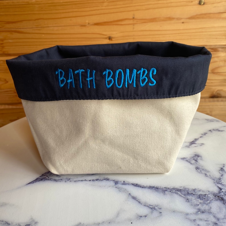 May include: A cream-coloured canvas bath bomb storage container with a navy blue fabric top.  The words 'BATH BOMBS' are embroidered in teal on the navy blue fabric. The container is shown on a marble surface.