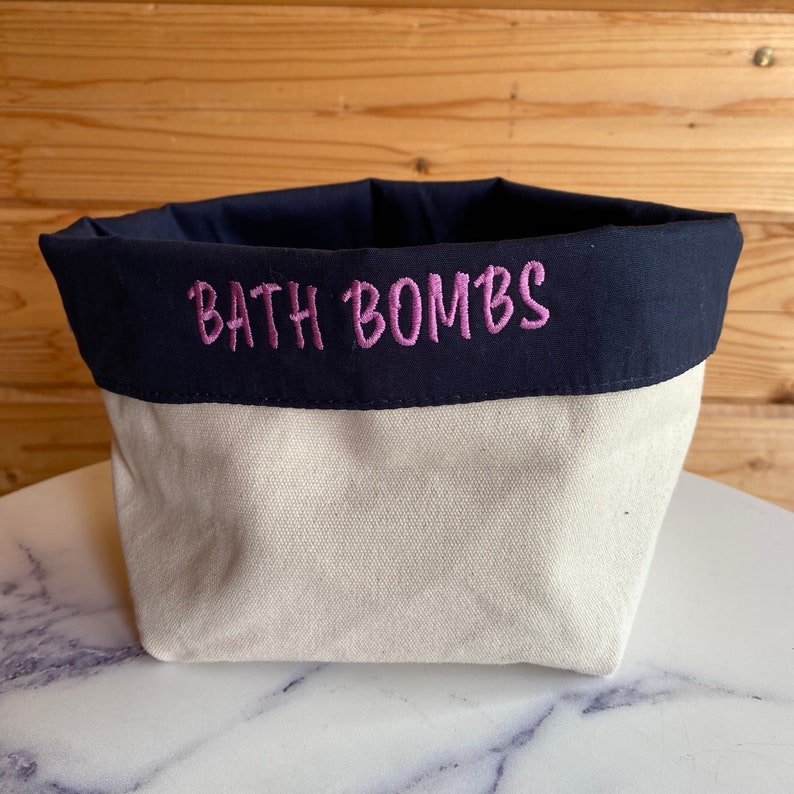 May include: A cream-coloured canvas bath bomb storage container with a navy blue fabric top.  The words 'BATH BOMBS' are embroidered in magenta on the navy blue fabric. The container is shown on a marble surface. The container is ideal for organizing and storing bath bombs.