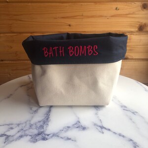 May include: A cream-coloured canvas bath bomb storage container with a navy blue top.  The words 'BATH BOMBS' are embroidered in red on the navy blue top. The container is sitting on a marble surface.