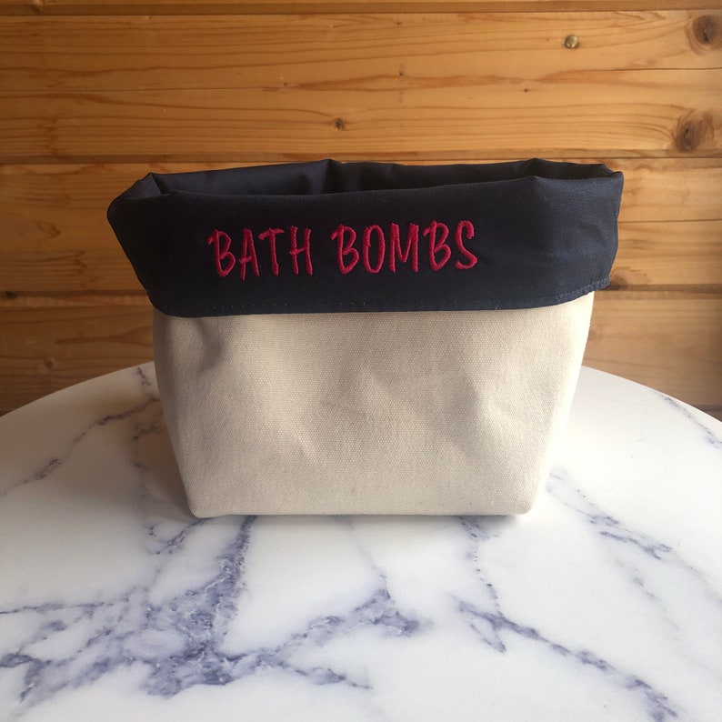 May include: A cream-coloured canvas bath bomb storage container with a navy blue top.  The words 'BATH BOMBS' are embroidered in red on the navy blue top. The container is sitting on a marble surface.