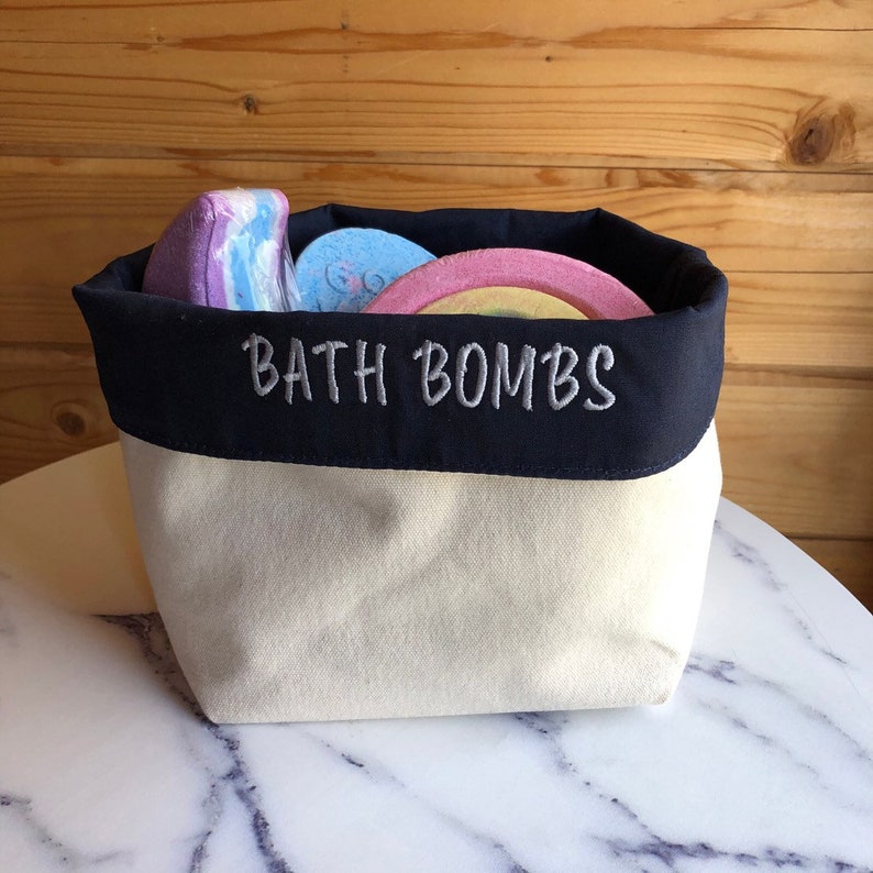 May include: A cream-colored canvas bath bomb storage container with a navy blue top. The navy blue top is embroidered with the text 'BATH BOMBS' in grey thread.  The container is filled with several colourful bath bombs in shades of purple, pink, light blue, and yellow. The bath bombs have a variety of shapes and designs.