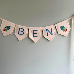 May include: A pink fabric banner with the name "BEN" spelled out in blue embroidery. Each letter is on a separate pennant with a rocket ship design on either end.