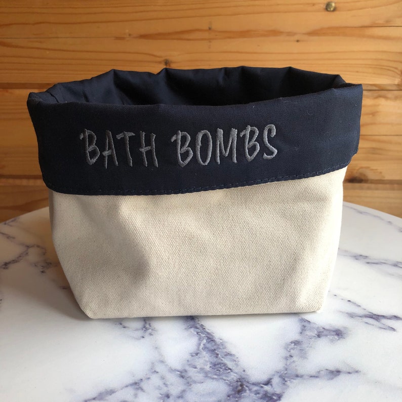 May include: A cream-coloured canvas bath bomb storage container with a navy blue fabric top. The words 'BATH BOMBS' are embroidered in grey on the navy blue fabric. The container is sitting on a marble surface.