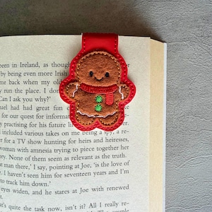 May include: A felt magnetic bookmark shaped like a gingerbread person with brown felt, a red scarf, and two green buttons. The gingerbread figure is outlined in red stitching and attached to a red rectangular magnetic backing.