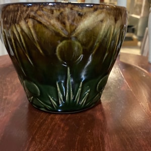 Puede incluir: Un cuenco de cerámica con un esmalte verde oscuro y marrón. El cuenco presenta un diseño floral con detalles en relieve. El cuenco está sobre una superficie de madera.