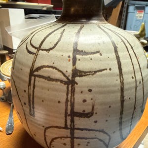 May include: A ceramic vase with a round body and a narrow neck. The vase is a light beige color with dark brown abstract lines and dots. The rim and neck are a darker brown. The vase is likely handmade.