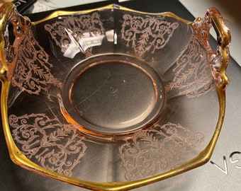 May include: A pink glass dish with gold trim and a delicate etched floral design. The dish has two handles and is ideal for serving snacks or desserts.