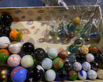 May include: A collection of colorful glass marbles in a variety of patterns and colors, including clear, blue, green, red, orange, yellow, and black. The marbles are in a box with a seashell pattern.