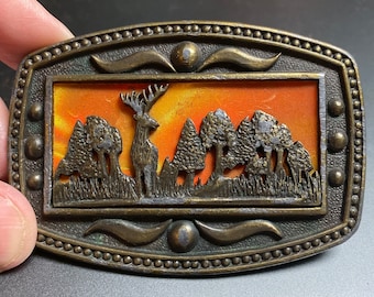 May include: A vintage belt buckle featuring a deer silhouette in a forest scene. The buckle is made of brass and has a textured finish. The scene is set against an orange sunset.