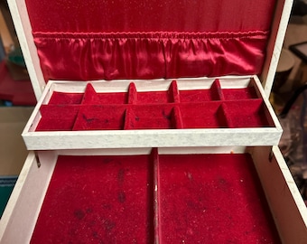 May include: Open, vintage jewelry box with a cream-colored exterior and a red velvet interior. The box has multiple compartments and hooks for hanging jewelry. The interior is lined with red satin and velvet.