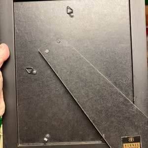 Puede incluir: La parte posterior de un marco de imagen negro con un gancho de suspensi&oacute;n plateado y una etiqueta que dice "Burnes of Boston".