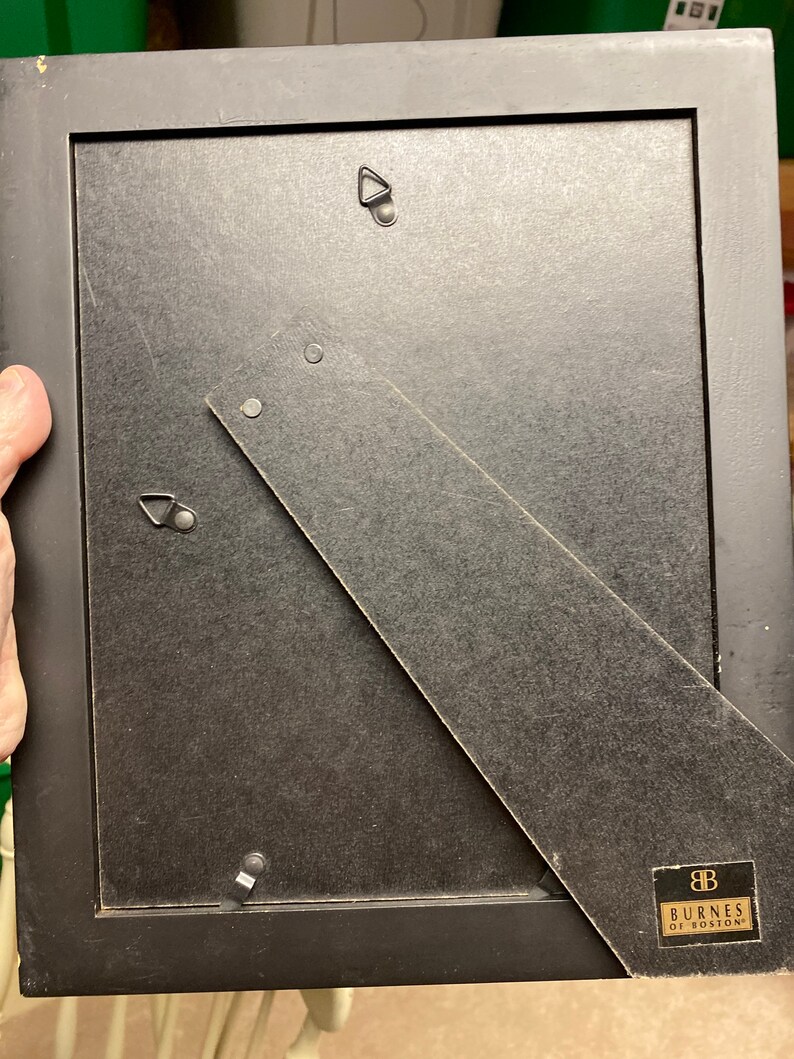 Puede incluir: La parte posterior de un marco de imagen negro con un gancho de suspensi&oacute;n plateado y una etiqueta que dice "Burnes of Boston".