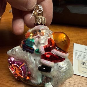 May include: A Christmas ornament featuring Santa Claus riding a polar bear. Santa wears a red suit with white trim and carries a gold sack. The polar bear is white with glitter accents, and Santa is holding a small train.