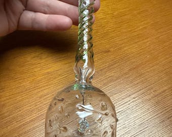 Könnte beinhalten: Eine durchsichtige Glasglocke mit einem grünen Spiralstiel und einem kleinen grünen Oberteil. Die Glocke hat eine strukturierte Oberfläche mit kleinen Blasen und einen ausgestellten Boden. Im Inneren hängt ein kleiner, klarer Kristall.