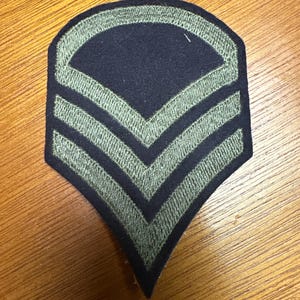 Könnte beinhalten: Dunkelblauer Militärpatch mit drei olivgrünen Chevrons. Der Patch hat eine gebogene Oberseite und eine spitz zulaufende Unterseite. Die Chevrons sind mit einem strukturierten Aussehen gestickt. Es ist wahrscheinlich ein Rangabzeichen.