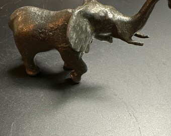 Puede incluir: Una pequeña figurita de elefante de metal gris con la trompa levantada en el aire. El elefante tiene una superficie texturizada y está de pie sobre una superficie gris.