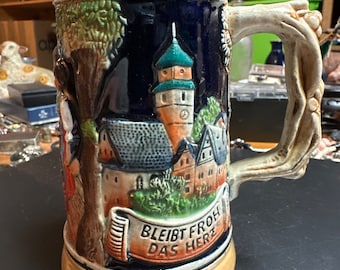 May include: A blue and brown ceramic beer stein featuring a scene of a town and a banner that reads "Bleibt froh das Herz".