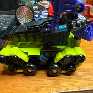 May include: A neon green and black toy tank with a purple cannon. The toy has black wheels and a black top section. The cannon is purple with a yellow lens. The toy is on a wooden surface.
