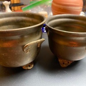 May include: Two antique brass pots with ornate handles and three small feet. The pots have a rounded shape and a tarnished finish, suggesting age and use. The handles are decorative, adding to the vintage aesthetic. These pots could be used for decorative purposes or as small planters.