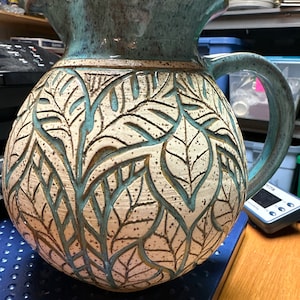 May include: A ceramic pitcher with a light blue glaze and a white body. The pitcher has a carved leaf design and a wide, flared lip.