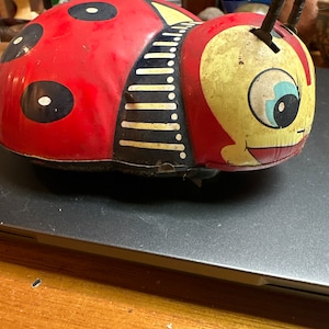 May include: Vintage ladybug-shaped tin toy with a red body, black spots, and a yellow head. The toy has a slot for coins and antennae. The ladybug has a smiling face and is likely a child's bank.