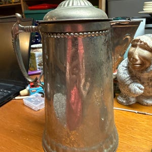 Könnte beinhalten: Eine Vintage-Kanne aus silberfarbenem Metall mit dekorativem Griff und Deckel. Die Kanne hat einen zylindrischen Körper mit einem Perlenbesatz und einer spitzen Spitze auf dem Deckel. Die Kanne wird wahrscheinlich zum Servieren von Getränken verwendet.