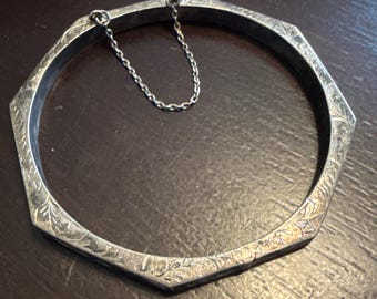 May include: A silver-toned octagonal bangle bracelet with a delicate chain. The bracelet features engraved floral patterns along its edges. The design is vintage and elegant, suitable for everyday wear or special occasions.
