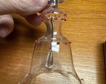 Könnte beinhalten: Klarer gläserner glockenförmiger Schmuck mit roten Akzenten. Der Schmuck hat oben eine Schlaufe zum Aufhängen und einen tropfenförmigen Kristallanhänger. Rote Perlen verzieren die Oberseite und den Rand der Glocke. Die Glocke wird von einer Hand gehalten.