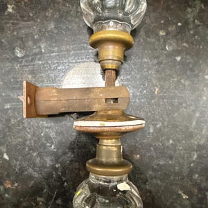 May include: Antique glass door knob set with brass hardware and a rusty metal bracket. The clear glass knobs have a faceted design. The brass hardware shows signs of age and wear. The set is designed for a door.