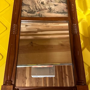 Puede incluir: Un espejo con marco de madera con una pequeña impresión de paisaje sobre un espejo más grande. El marco tiene un diseño tallado decorativo.