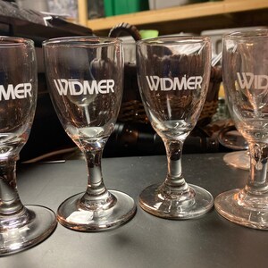 Puede incluir: Cuatro copas de licor transparentes con la palabra "WIDMER" impresa en blanco. Las copas tienen una base redondeada, un tallo delgado y un borde acampanado. Las copas están dispuestas en fila sobre una superficie oscura.