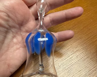 Könnte beinhalten: Eine transparente Glasglocke mit blauem Blumendesign. Die Glocke hat einen langen, schlanken Griff und einen ausgestellten Sockel. Das blaue Design befindet sich am unteren Teil der Glocke. Die Glocke wird in einer Hand gehalten.