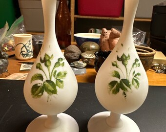 Puede incluir: Dos jarrones de vidrio esmerilado blanco con base bulbosa y parte superior acampanada. Cada jarrón presenta un diseño pintado a mano de hojas y flores verdes. Los jarrones se exhiben sobre una superficie oscura.