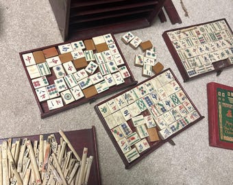 May include: A wooden box with a brass handle, containing a set of mahjong tiles. The tiles are arranged in a grid pattern, with some tiles scattered around the box. The box is open, revealing the tiles inside. There is also a book titled "The Famous Chinese Game of Mahjong" visible in the image.