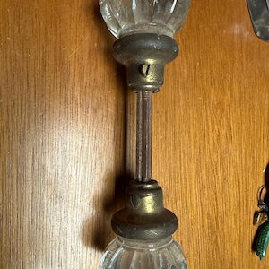 May include: A vintage door knob set featuring two clear, fluted glass knobs attached to a central metal spindle. The knobs are secured by tarnished brass-toned metal collars, each featuring a small flat-head screw for adjustment and mounting.