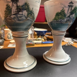 May include: Two white ceramic cups with a blue and green lighthouse scene painted on the side. The cups have a white base with a blue rim.