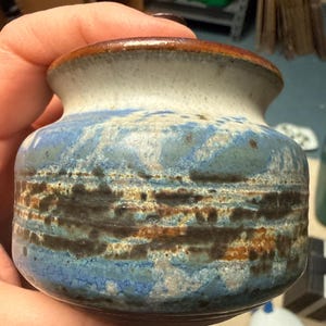 May include: A small, handcrafted ceramic jar with a wide base and a narrow opening. The jar features a textured surface with a blend of blue, brown, and white glaze. The rim is a dark brown color.