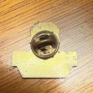 May include: A vintage gold-toned metal pin with a clasp on the back. The pin has a rectangular base with a rounded top and a detailed design. The pin is set against a wooden surface.