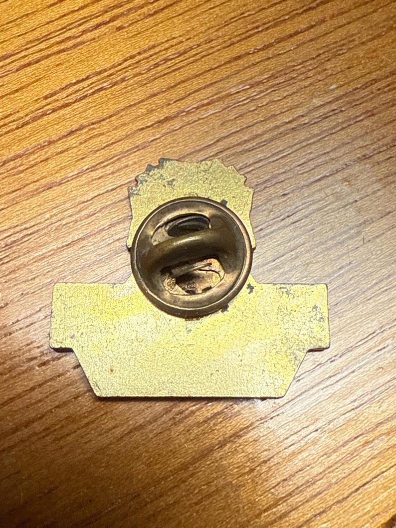 May include: A vintage gold-toned metal pin with a clasp on the back. The pin has a rectangular base with a rounded top and a detailed design. The pin is set against a wooden surface.