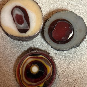 May include: Three decorative, circular objects with unique, layered designs. One features a cream-colored outer ring with a dark red and white center. Another has a gray outer ring with a red and white center. The third has concentric rings of red, yellow, and black.