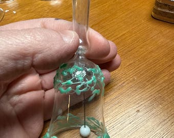 Könnte beinhalten: Eine transparente Glasglocke mit langem Griff, verziert mit hellgrünen, rankenartigen Designs. Im Inneren der Glocke hängt eine kleine weiße Kugel. Die Glocke wird in einer Hand gehalten, weitere Glocken sind im Hintergrund zu sehen.