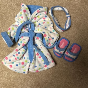 Puede incluir: Una bata de felpa blanca con ribete azul y lunares de colores, una diadema a juego y un par de zapatillas rosas y azules. La bata tiene cinturón y capucha. Las zapatillas tienen la palabra "American".