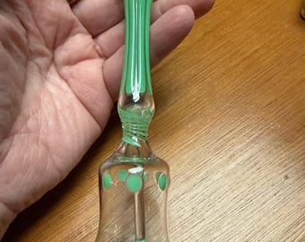 Könnte beinhalten: Eine transparente Glasglocke mit grünem Griff und Akzenten. Der Griff ist mit vertikalen grünen Streifen verziert, und die Glocke hat grüne Tupfen. Die Glocke wird in einer Hand gehalten.