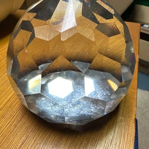 Op de afbeelding: Een heldere, facetgeslepen kristallen presse-papier met een geometrisch ontwerp. De bolvormige presse-papier heeft meerdere platte oppervlakken die licht reflecteren. Het rust op een houten oppervlak, wat de helderheid en vorm benadrukt. Een decoratief bureau-accessoire.
