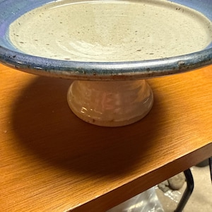 May include: A ceramic pedestal bowl featuring a wide, shallow basin with a speckled off-white interior and a deep blue glazed rim. The bowl is supported by a sturdy, flared base that matches the off-white speckled finish of the basin interior.