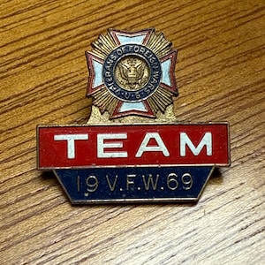 May include: A vintage metal pin with the text "TEAM" in white letters on a red background. Below, it reads "19 V.F.W. 69" in white on a blue background. The top features a gold emblem with "VETERANS OF FOREIGN WARS U.S." in a circular design.