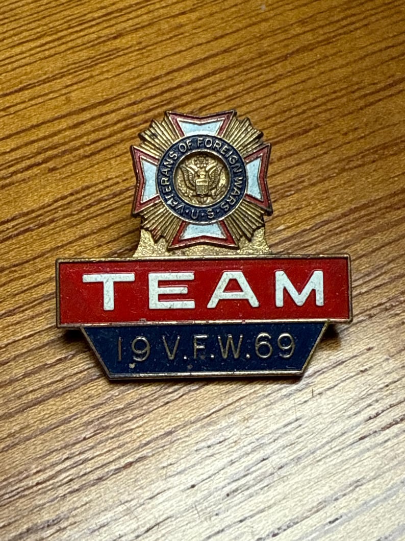 May include: A vintage metal pin with the text "TEAM" in white letters on a red background. Below, it reads "19 V.F.W. 69" in white on a blue background. The top features a gold emblem with "VETERANS OF FOREIGN WARS U.S." in a circular design.