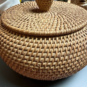 May include: A round, woven wicker basket with a lid and a knob on top. The basket is made of natural brown wicker and has a smooth, even weave.