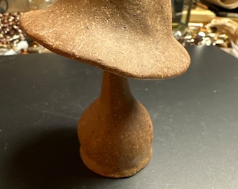May include: A brown ceramic mushroom sculpture with a wide, rounded cap and a slender stem. The sculpture has a rough, textured surface.
