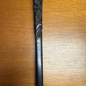 May include: A dark, carved wooden handle with floral and vine details. The handle is rectangular with a faceted top and a smooth, polished finish. The carving is intricate and detailed, showcasing the craftsmanship of the piece.